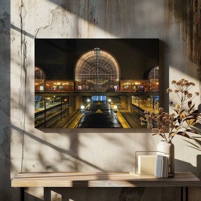 A symmetrical view of a modern train station at night, with a large illuminated arched glass window. Below, empty, wet train tracks and platforms reflect the station's lights, while cars cross an overpass in the middle ground. Wall Art