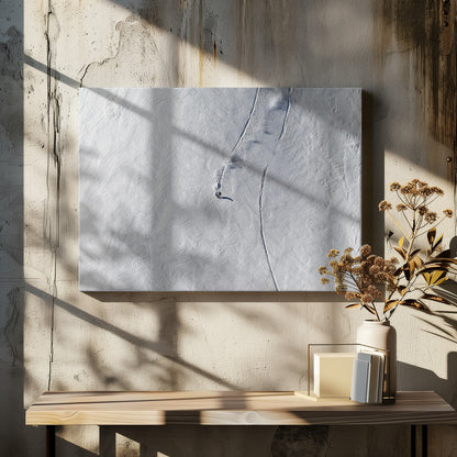 An aerial, top-down photograph of a lone snowboarder carving a turn down a vast, textured, snowy mountain slope, leaving fresh tracks in the powder. The image is presented in a simple, grey frame. Wall Art