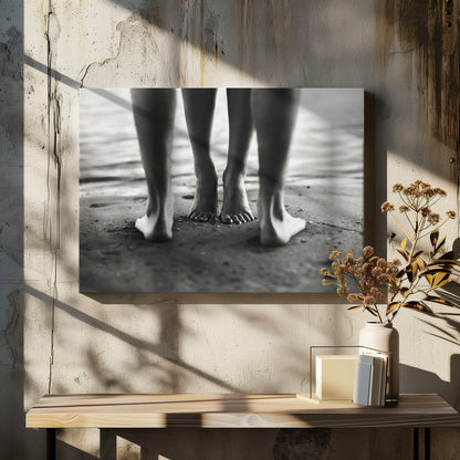 A framed, black and white photograph showing a low-angle view of two people's bare feet and lower legs standing on a wet sandy beach. The central pair of feet is in focus, while another person's legs in the foreground are blurred, creating depth. The water's edge is visible in the background. Artwork