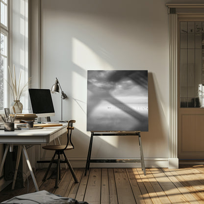A minimalist black and white photograph in a light wood frame, depicting a calm, serene lake under a heavy, cloudy sky. The still water reflects the sky, with a row of weathered wooden posts breaking the surface along the horizon. Print