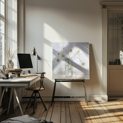 A delicate still life photograph of several white daisies with bright yellow centers in a clear glass jar. The arrangement sits on a light wooden surface against a backdrop of soft, draped white fabric. The entire image is enclosed in a light wood frame. Artwork
