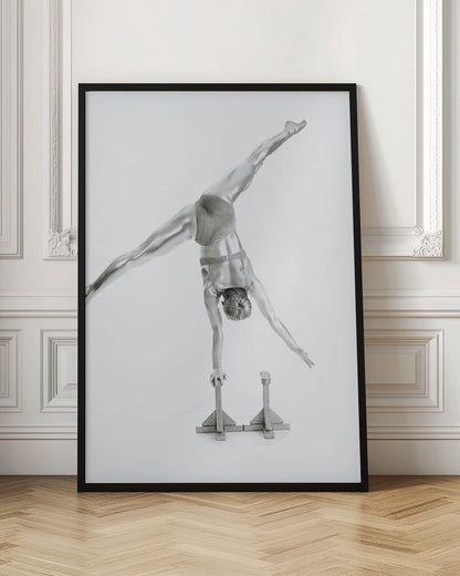 A black and white studio photograph of a muscular gymnast coated in silver paint, performing a dynamic one-armed handstand on a small block. Their legs are split wide apart in a display of strength and flexibility against a clean white background, all enclosed in a black frame. Decor