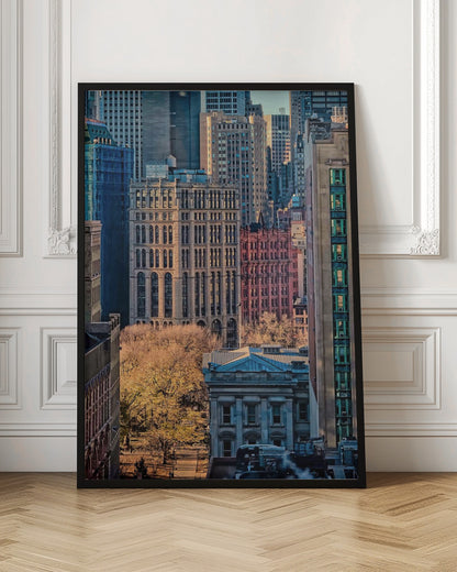 A high-angle view of a dense city skyline featuring a mix of old and new architectural styles. Golden light illuminates the autumn foliage of a park and the facades of several buildings, creating a warm contrast with the cool blue tones of the skyscrapers in the background. Decor