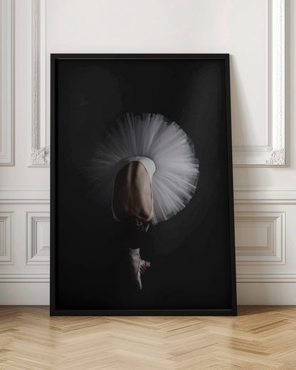 A dramatic photograph of a ballerina against a solid black background. She is bent completely forward, showing her arched back to the camera. Her white tutu fans out in a wide circle around her, and one foot in a pointe shoe is visible beneath her, balanced on its tip. Print