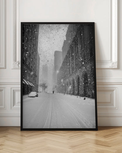 A grainy, black and white photograph captures a deserted city street during a heavy snowstorm. Tall buildings line the street, which is covered in a thick blanket of snow. A single car on the left is almost completely buried, and in the distance, a lone figure walks with an umbrella down the center of the empty road. Poster