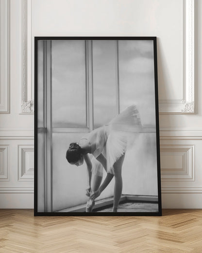 A black and white photograph of a ballerina in a white tutu, bending over to tie her pointe shoe in front of a large, bright window. The motion of her tutu is captured in a graceful blur. Print