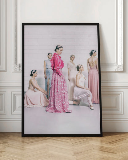 A studio photograph of six young women in elegant pastel gowns posing against a white brick wall. The central figure wears a vibrant pink ruffled blouse and a patterned maxi skirt, while the others are dressed in light pink, white, and pale blue dresses, some with ballet-inspired elements. They all have flowers in their hair, creating a soft, high-fashion aesthetic. Print
