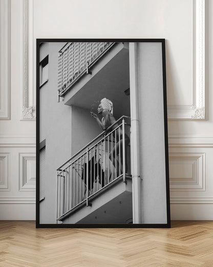 A black and white photo of a woman with a towel on her head, smoking a cigarette on an apartment balcony. The low-angle shot captures her leaning on the metal railing, with smoke drifting from the cigarette. Print