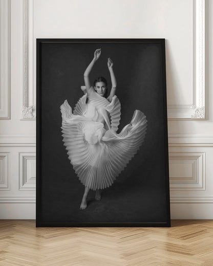 A framed, black and white photograph of a woman in a dramatic pose, wearing a voluminous white pleated dress. Her arms are raised above her head as the dress fans out around her in a sculptural, swirling motion. She looks directly at the camera against a solid dark gray background. Decor