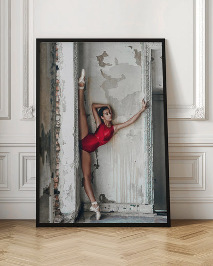 A ballerina in a shiny red leotard performs a standing split against a distressed, peeling wall. Her left leg is extended straight up, and her body leans against the wall in a display of incredible flexibility and grace amidst a rustic, derelict setting. Artwork