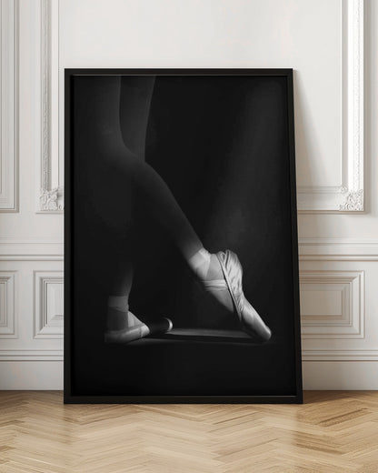 A dramatic black and white photograph showing the lower legs and feet of a ballet dancer, with one foot gracefully en pointe, highlighted by strong side lighting against a deep black background. Decor