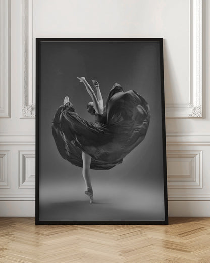 A dramatic black and white photograph of a ballerina captured mid-twirl. She is perfectly balanced on the tip of one pointe shoe, with her other leg kicked high. Her arms are raised elegantly above her head, and her long, dark dress billows out around her in a magnificent, fluid circle against a solid gray background. Wall Art
