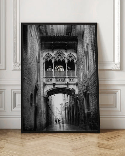 A moody, vertical black and white photograph of an ornate Gothic-style bridge connecting two stone buildings over a narrow alleyway. Below the arch of the bridge, the wet street stretches into the distance where two figures are walking away. Decor