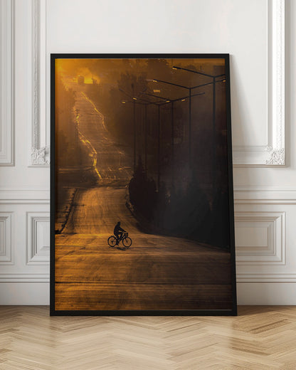 A silhouette of a person riding a bicycle across a wide, winding road illuminated by the warm, golden light of a sunrise or sunset. The road stretches uphill into the distance, and tall streetlights stand in shadow on the right. Artwork