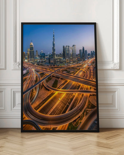 A long-exposure, aerial photograph of the Dubai skyline at dusk. The iconic Burj Khalifa stands tall in the center, surrounded by other illuminated skyscrapers against a deep blue twilight sky. In the foreground, a complex web of highways and overpasses glows with the orange and white light trails of moving traffic. Decor