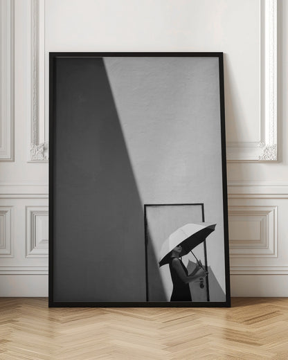 A minimalist black and white photograph of a woman in a black dress holding a two-toned umbrella. She stands in front of a door against a light wall, with a sharp, diagonal shadow dramatically cutting across the upper left of the image, creating a high-contrast, geometric composition. Decor
