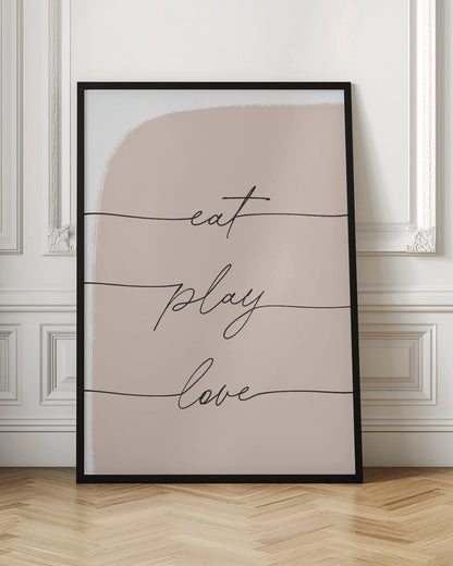 A modern typographic art print in a black frame. The words 'eat', 'play', and 'love' are written in a black cursive script, each on a separate line against a pale pink painted background. Print
