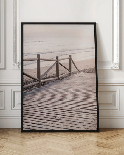 A wooden boardwalk with a rustic log fence leads down to a sandy beach with gentle waves lapping at the shore under a soft, hazy sunrise or sunset. Decor