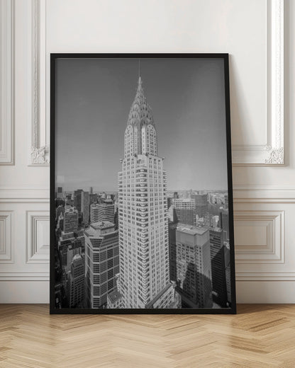 A high-angle, black and white photograph of the iconic Chrysler Building, its Art Deco spire prominently featured against the New York City skyline under a clear sky. Artwork