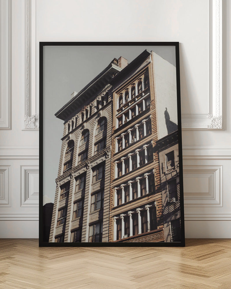 A low-angle, sunlit photograph of two ornate, multi-story historic buildings against a pale sky, framed in black. The detailed facades feature arched windows and decorative columns, with warm light creating strong shadows. Poster