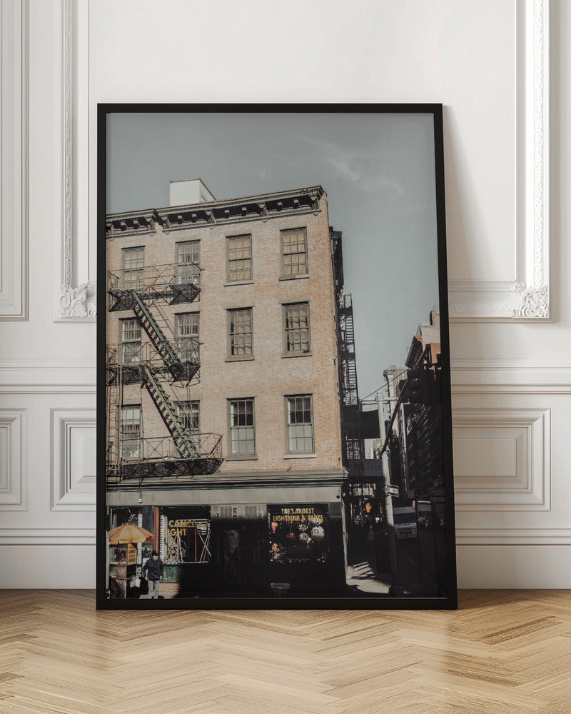 A low-angle, desaturated photograph of a classic brown brick building in a city, featuring multiple windows and a black metal fire escape zigzagging up the side. The sky is a pale, hazy blue, and the ground floor has dark storefronts. Decor