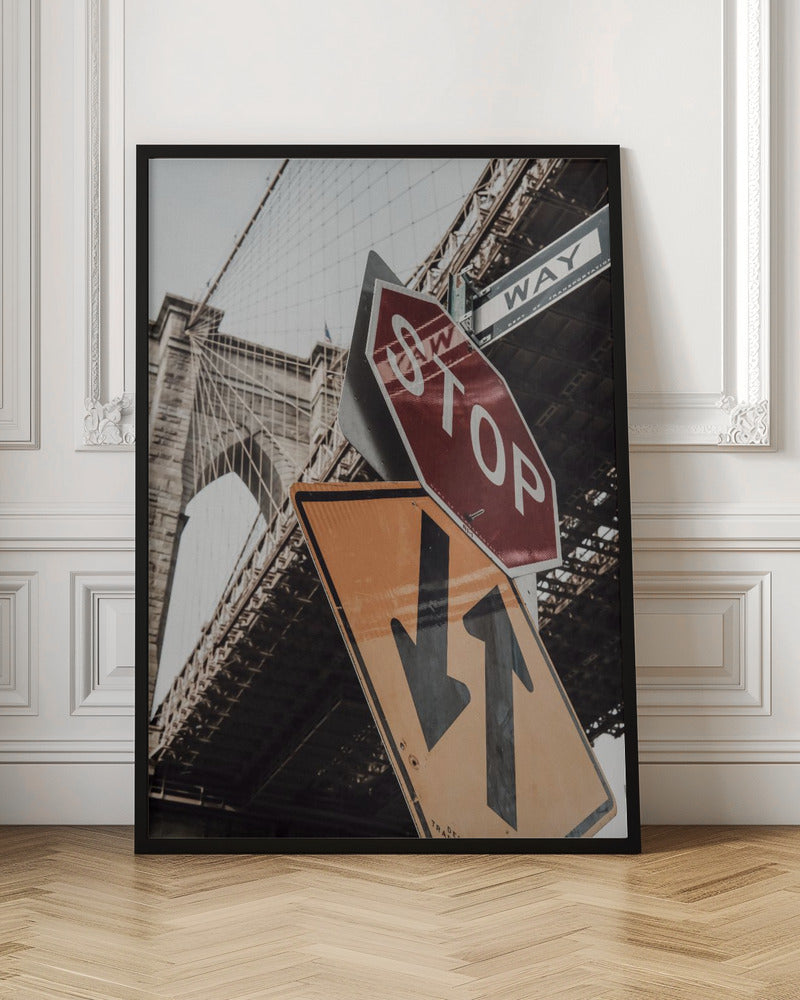 A low-angle photograph of the Brooklyn Bridge with a collection of street signs in the foreground. A red stop sign and a yellow sign with two arrows are prominent against the architectural details of the bridge. Print