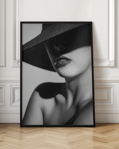 A dramatic black and white close-up photograph of a woman wearing a large, dark, wide-brimmed hat. The hat casts a deep shadow over the upper half of her face, obscuring her eyes and creating a sense of mystery. Only her nose, full lips, chin, neck, and shoulder are visible, highlighted by a strong light source. Decor