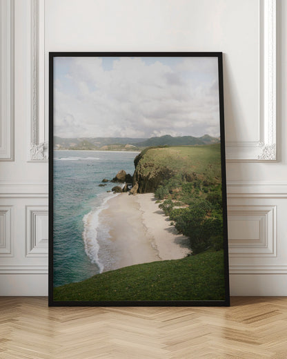 A high-angle photograph of a secluded tropical beach, with turquoise waves gently washing onto the white sand. A lush green cliff covered in grass and small trees frames the cove, with distant rolling hills visible under a soft, cloudy sky. Decor
