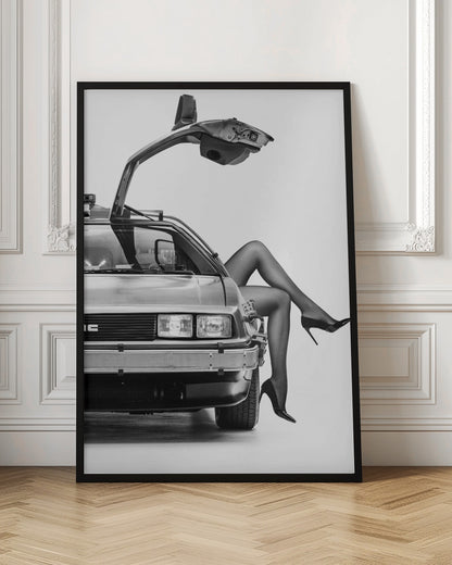 A black and white studio photograph of a DeLorean car with its gull-wing door open, featuring a woman's legs in black stockings and stilettos elegantly emerging from the passenger side. Decor