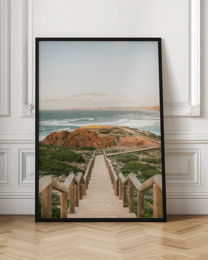 A wooden staircase and walkway descends a green, scrub-covered hill towards a rocky point overlooking a churning blue ocean under a pale, hazy sky. Print