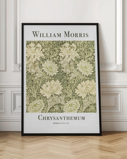 A framed art print of William Morris's 'Chrysanthemum' pattern from 1877. The design features a dense, repeating pattern of large, cream-colored chrysanthemums and foliage in shades of sage green on a dark green background. The poster has a white border with the artist's name at the top and the title at the bottom. Print