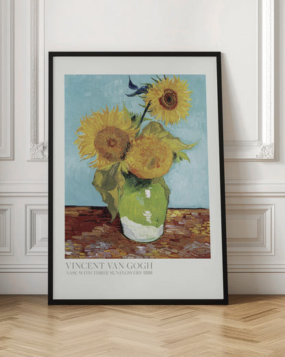 A framed art print of Vincent van Gogh's painting 'Vase with Three Sunflowers' from 1888. The artwork features three large, vibrant yellow sunflowers in a green vase set against a textured light blue background. The painting is in the post-impressionist style with thick, visible brushstrokes. The print has a white border with text at the bottom identifying the artist and title. Print