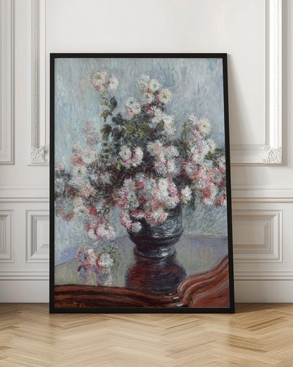 An impressionist painting by Claude Monet depicting a large bouquet of white and pink chrysanthemums in a dark, ornate vase. The vase sits on a reflective, reddish-brown wooden table, and the background is a textured light blue wall. The painting is framed in black. Artwork