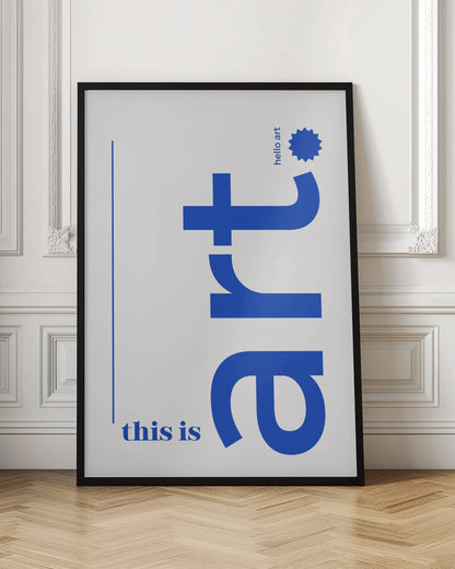 A minimalist art print in a black frame, featuring a white background with blue text. The word 'art' is written vertically in a large, bold lowercase font. To the left, smaller text reads 'this is'. At the top right, 'hello art' is written next to a blue starburst graphic. Wall Art