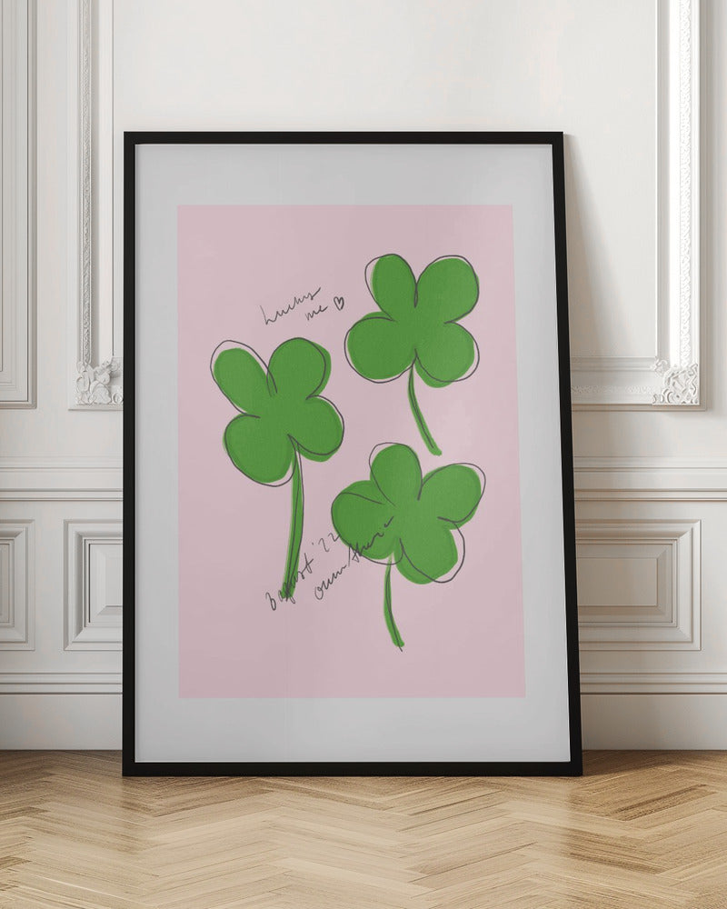 A framed art print featuring three hand-drawn four-leaf clovers in green with a loose black outline, set against a solid light pink background. Cursive handwriting on the print reads 'lucky me ♡' and includes a signature. Artwork