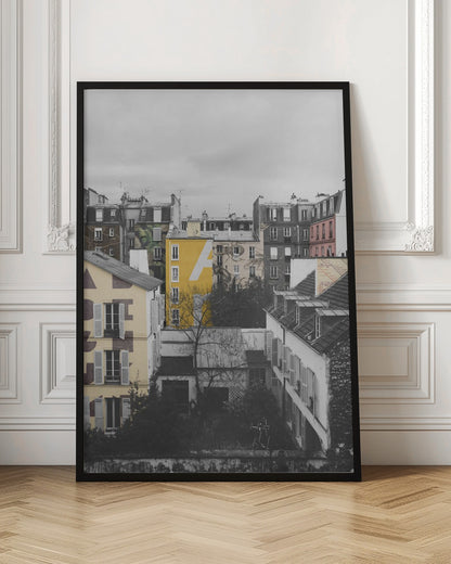 A framed, vertical photograph with selective color, showing a dense cluster of Parisian-style buildings under an overcast sky. Most of the image is in black and white, but one building is a vibrant yellow with a large white 'A' painted on it, creating a striking focal point. Wall Art