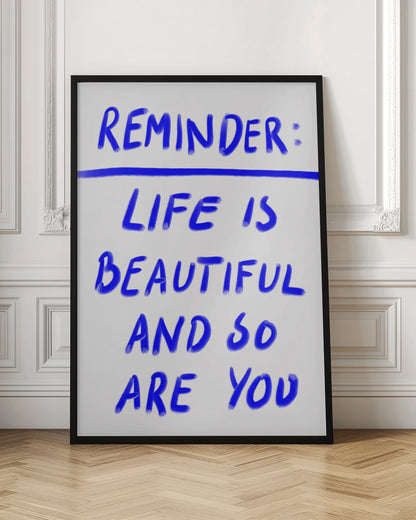 A framed poster featuring a positive affirmation handwritten in a blue, brush-stroke style font on a white background. The text reads 'REMINDER: LIFE IS BEAUTIFUL AND SO ARE YOU', with a blue line separating 'REMINDER:' from the rest of the quote. Print