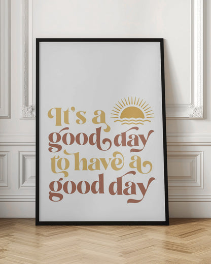 A framed art print with a white background featuring the quote 'It's a good day to have a good day' in a retro, curvy font. The text color alternates between terracotta brown and mustard yellow. Above the text is a small yellow icon of a sun rising over waves. Wall Art