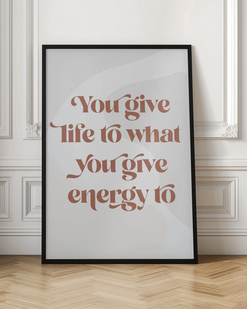 A framed motivational poster featuring the quote 'You give life to what you give energy to'. The text is printed in a dusty rose color using a stylish, retro serif font on a light gray background with subtle wavy shapes. Poster