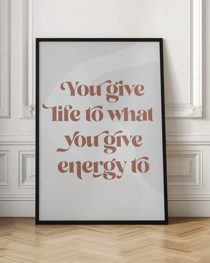 A framed motivational poster featuring the quote 'You give life to what you give energy to'. The text is printed in a dusty rose color using a stylish, retro serif font on a light gray background with subtle wavy shapes. Poster