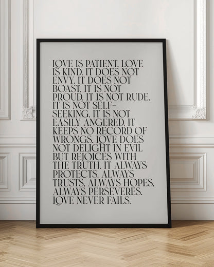 A minimalist art print featuring the 'Love is patient, love is kind...' bible verse from 1 Corinthians 13. The text is printed in a black, all-caps serif font on an off-white background and is surrounded by a simple black frame. Print
