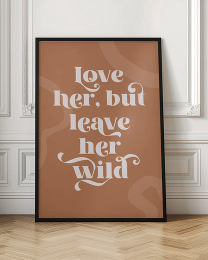 A framed typography poster with a terracotta brown background. In the center, the quote 'love her, but leave her wild' is written in a stylized, decorative, cream-colored font. Faint, lighter brown abstract shapes are visible in the background. Decor