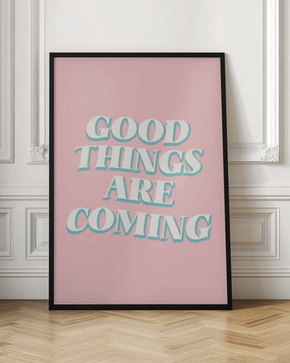 A framed poster with a light pink background featuring the motivational phrase 'GOOD THINGS ARE COMING'. The text is in a white, retro-style serif font with a light blue drop shadow, giving it a three-dimensional effect. Poster