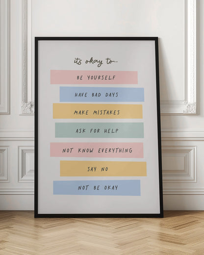 A framed inspirational poster with a white background. At the top, in a black handwritten font, it says 'it's okay to...'. Below is a list of affirmations, each on a different colored pastel rectangle: 'BE YOURSELF' on pink, 'HAVE BAD DAYS' on blue, 'MAKE MISTAKES' on yellow, 'ASK FOR HELP' on green, 'NOT KNOW EVERYTHING' on pink, 'SAY NO' on yellow, and 'NOT BE OKAY' on blue. Poster