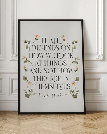 A framed art print featuring an inspirational quote by Carl Jung. The text reads 'It all depends on how we look at things, and not how they are in themselves' in a black serif font against an off-white background. The quote is encircled by a delicate wreath of watercolor botanicals with green leaves and pale orange flowers. Poster