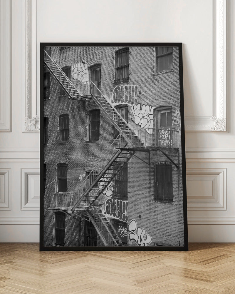 A black and white photograph of a brick apartment building with a prominent metal fire escape zigzagging across its facade. The walls are adorned with various styles of graffiti, and the windows are covered with bars, creating a gritty, urban scene. Decor