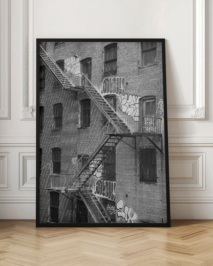 A black and white photograph of a brick apartment building with a prominent metal fire escape zigzagging across its facade. The walls are adorned with various styles of graffiti, and the windows are covered with bars, creating a gritty, urban scene. Decor