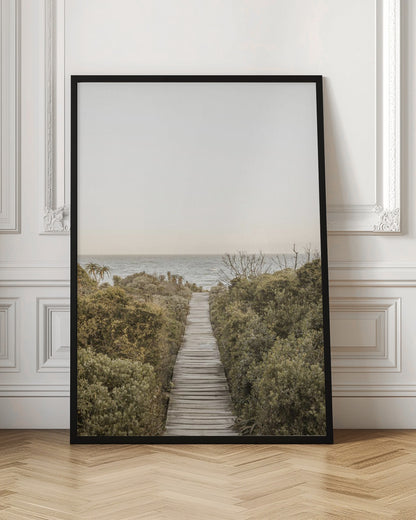 A vertical photograph of a weathered wooden boardwalk leading through dense coastal greenery towards the ocean under a pale, hazy sky, framed in black. Wall Art