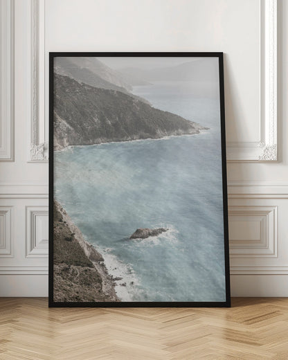 A high-angle photograph of a rugged, green coastline meeting a misty, light-blue ocean. The hazy atmosphere softens the view of the distant mountains and the waves crashing against the rocky shores. Poster