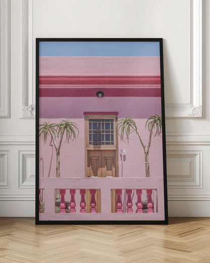 A brightly colored pink building stands under a clear blue sky, framed in black. A central wooden door with a paned window is flanked by two slender, palm-like trees. In the foreground, a pink balcony wall features fuchsia-colored balusters. Decor